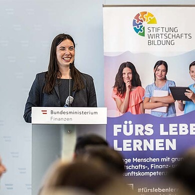 Frau spricht bei Veranstaltung über Finanzbildung an Schulen, im Hintergrund Stiftung Wirtschaftsbildung.