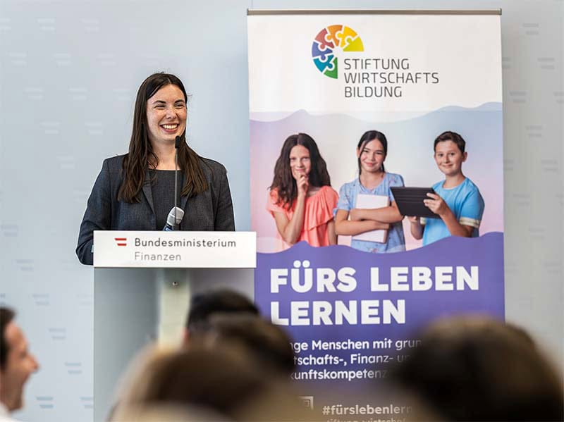 Frau spricht bei Veranstaltung über Finanzbildung an Schulen, im Hintergrund Stiftung Wirtschaftsbildung.